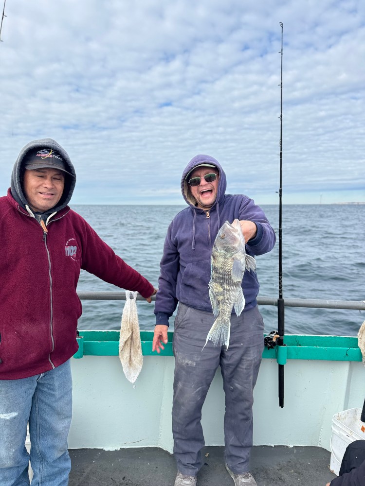Two people on a boat; one holds a fish, both wearing hoodies, with fishing rod and ocean in background.
