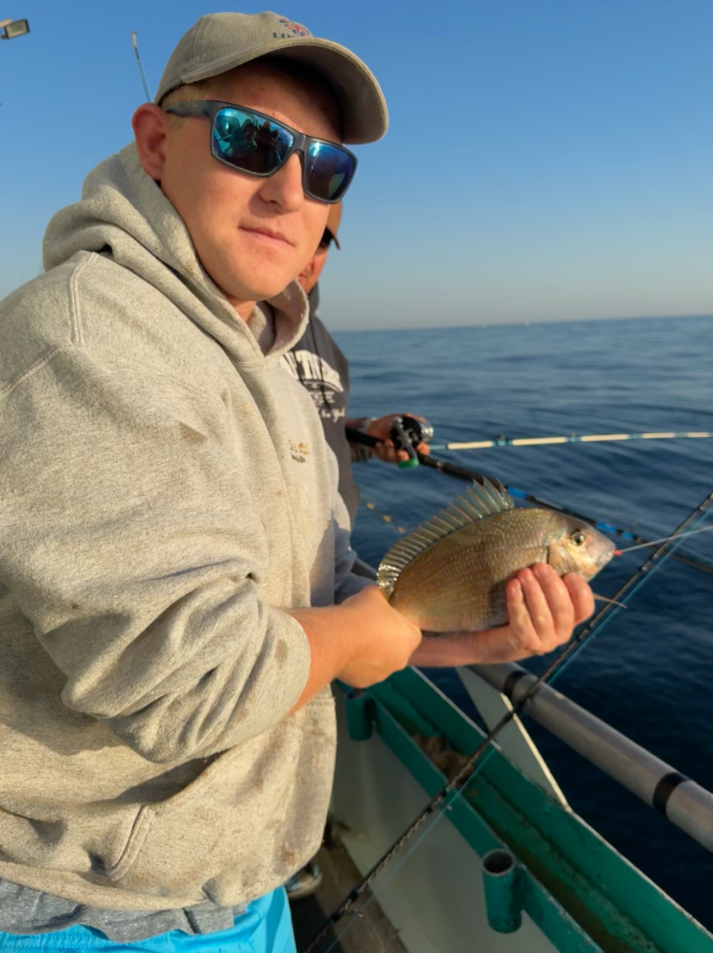 Person wearing sunglasses and a hoodie holding a fish on a boat.