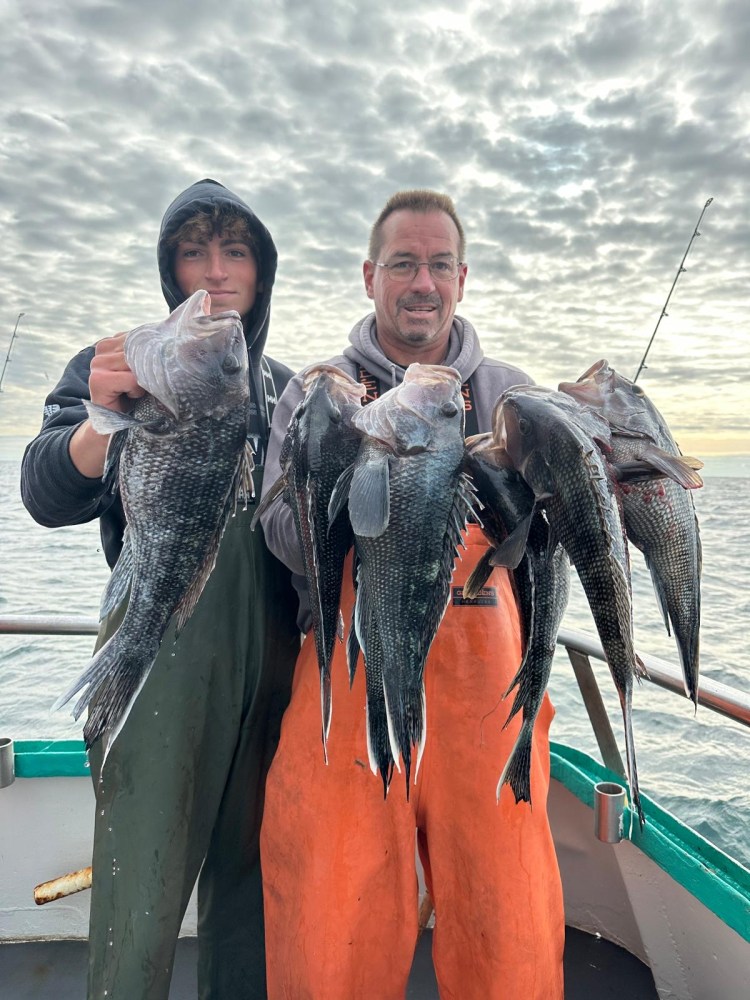 Two people on a boat holding several large fish with a cloudy sky backdrop.