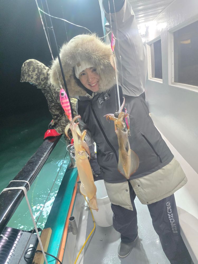 Person in winter clothing holding two squid on a fishing boat at night.
