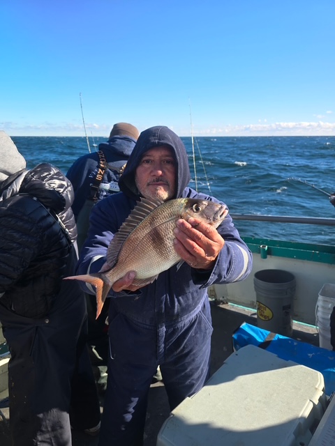 Person in a hooded jacket holding a fish on a boat with an ocean backdrop.