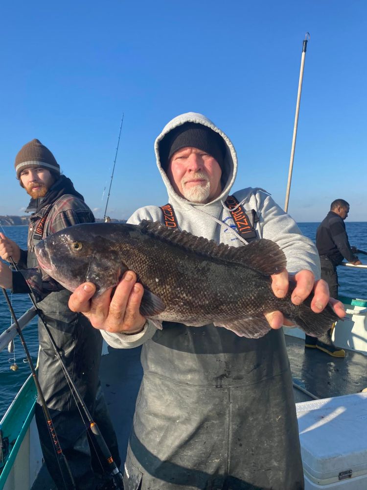 Man on boat holding large fish, with others fishing in background under a clear blue sky.