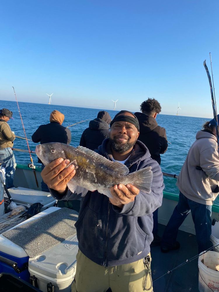 Man on boat holds fish, with wind turbines and ocean in the background.