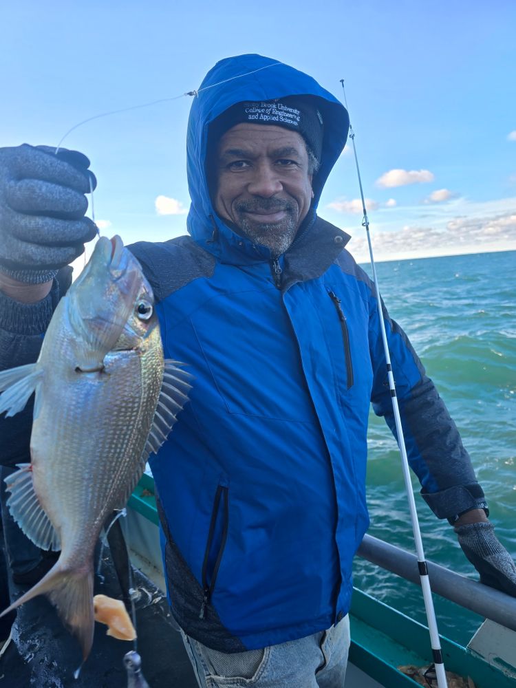 Man in blue jacket holding a fish on a boat with a fishing rod.