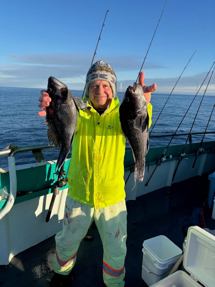 Person on a boat holding two fish, wearing a yellow jacket and knit hat, with fishing rods in the background.