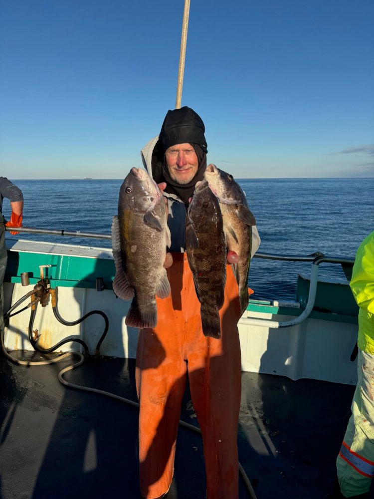 Person in orange overalls on boat holding two large fish against clear blue sky.