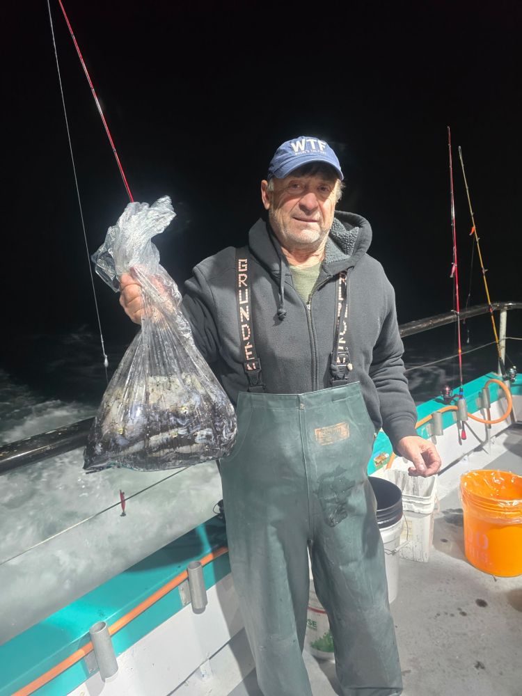 Man in green overalls holding a bag of fish on a boat at night.