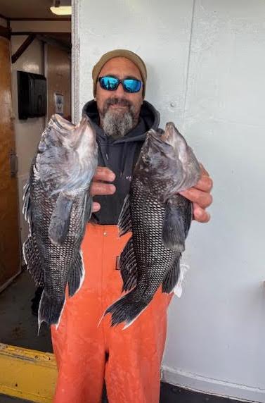 Person in sunglasses holds two large fish on a boat, wearing orange overalls and a brown beanie.