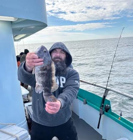 Person with a beard on a boat holding a fish in both hands, with a fishing rod in the background.