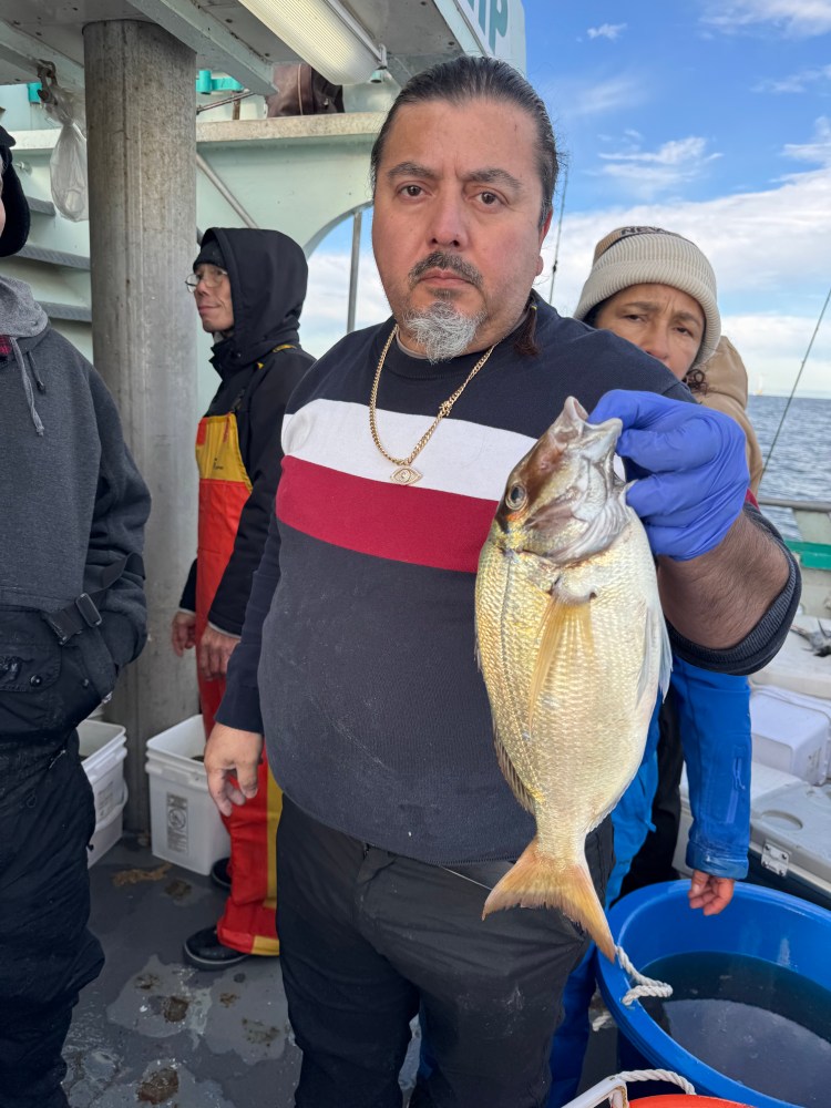 Man on boat holding a large fish, wearing blue gloves and a striped sweater, with others in the background.