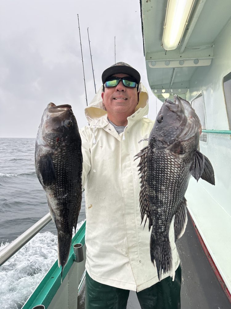 Person in white coat holding two large fish on a boat with ocean in the background.