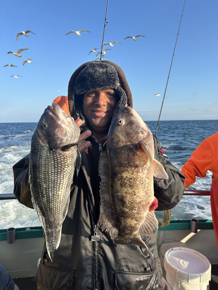 Man on boat holding two large fish, with seagulls flying overhead.