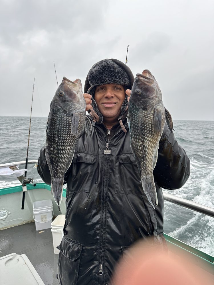 Person in winter coat holding two fish on a boat with ocean backdrop.