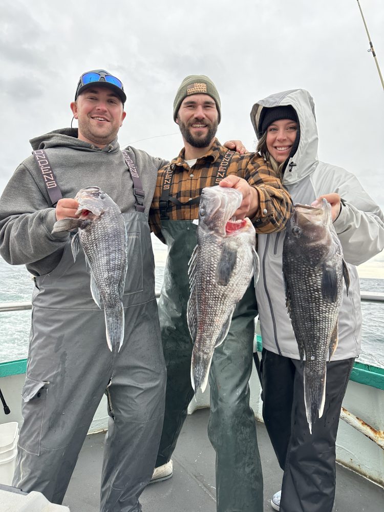 Three people on a boat holding large fish, wearing winter clothing.
