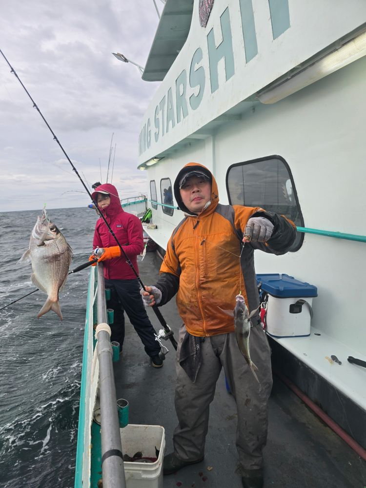 Two people on a boat holding fishing rods with fish caught, one wearing an orange jacket.