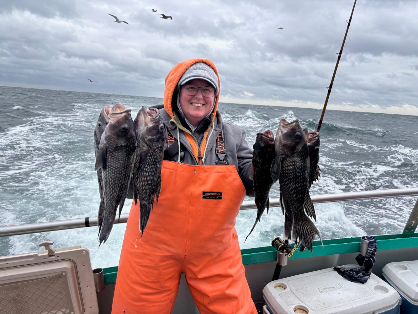 Person in orange overalls holding several fish on a boat with ocean and cloudy sky background.