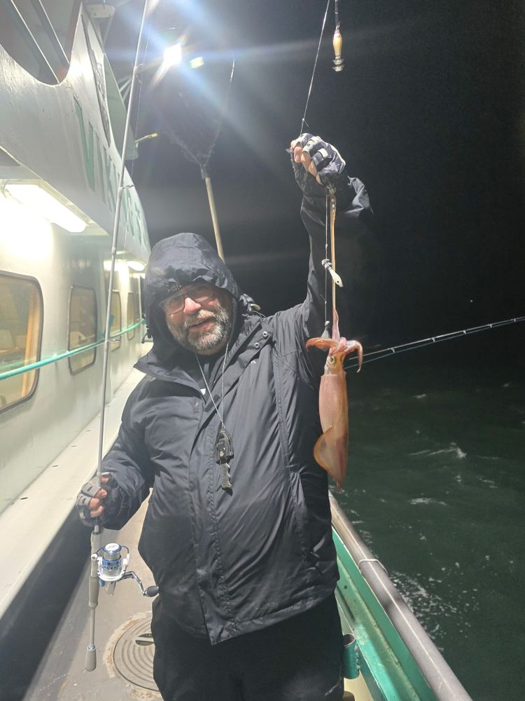 Person in hooded jacket holding a squid on a fishing line at night on a boat.