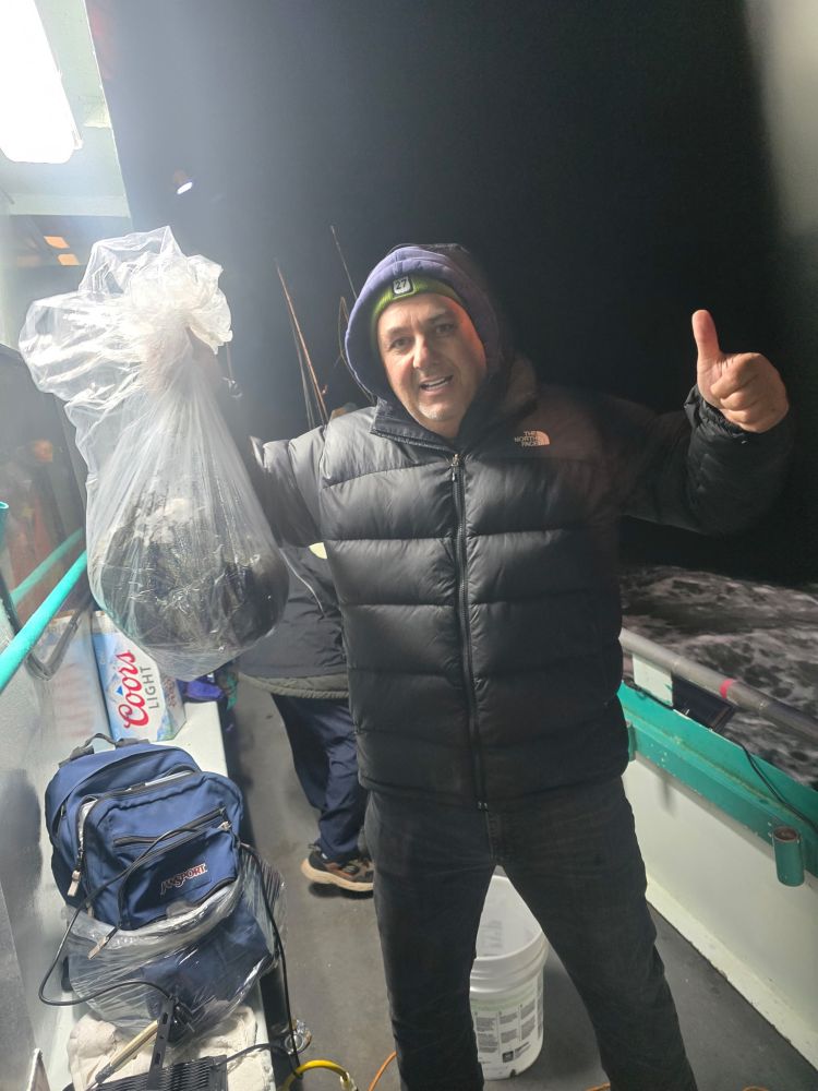 Person in a puffy jacket holds a bag and gives a thumbs-up on a boat at night.
