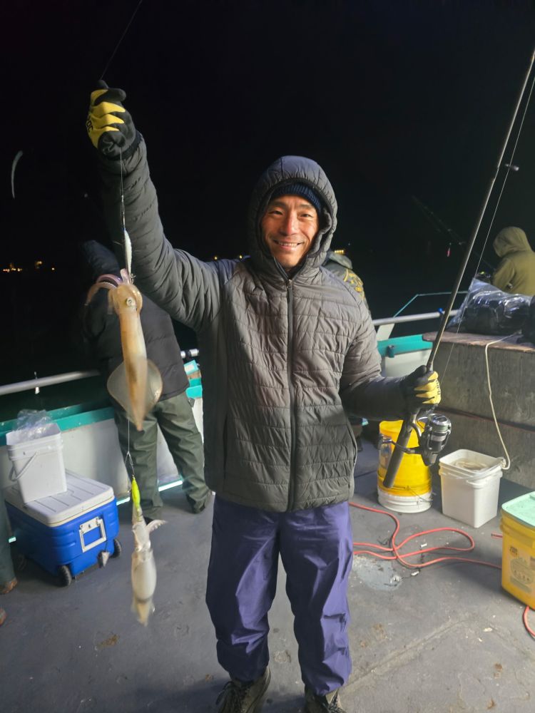 Person in winter clothing holding a fishing rod with a caught squid on a boat at night.