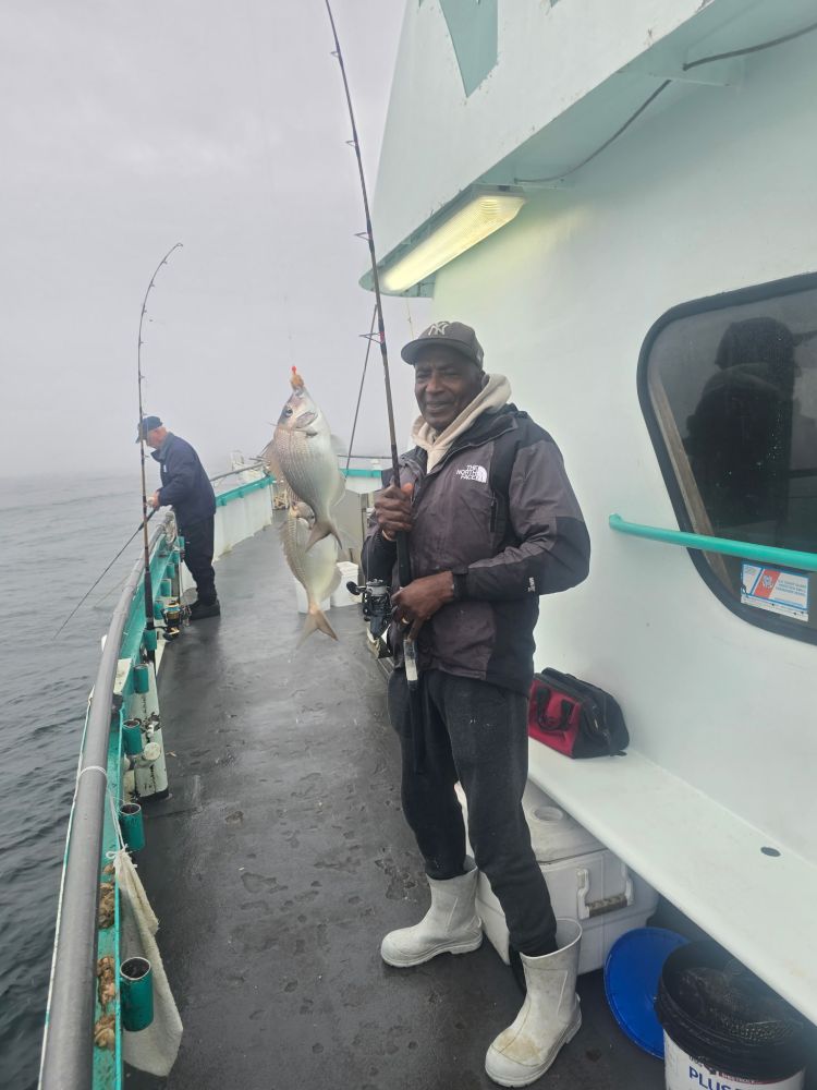 Man on boat holding fishing rod with large fish, another man fishing in background.