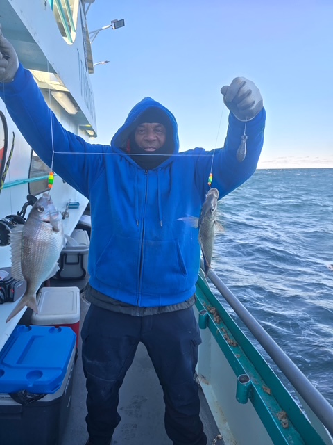 Person in blue hoodie holding two fish on a boat with ocean in background.