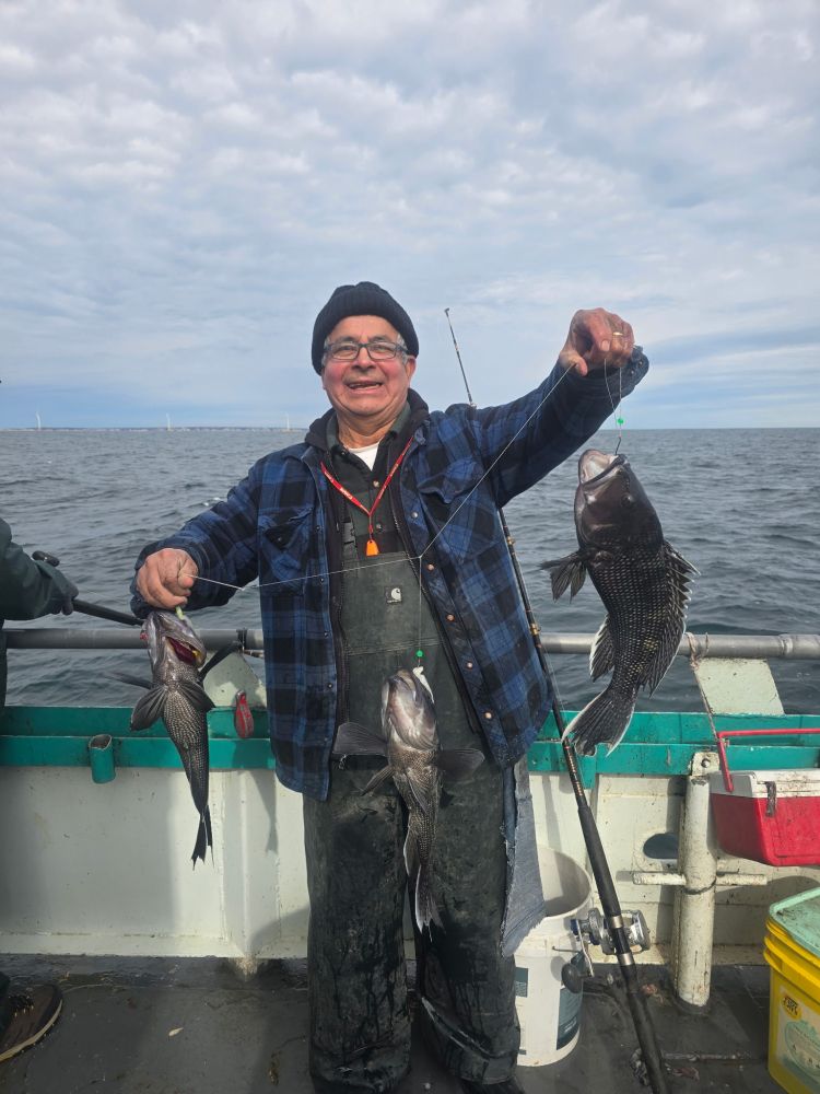 Man in a plaid shirt on a boat holding up three fish against a cloudy sky.
