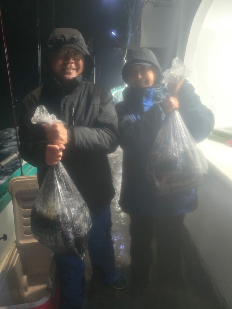 Two people on a boat holding bags of fish, wearing jackets and hoods at night.