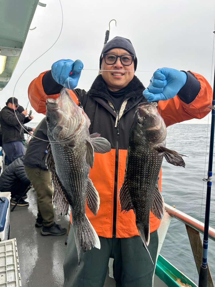 Person on a boat holding two large fish with gloved hands, wearing an orange and black jacket.
