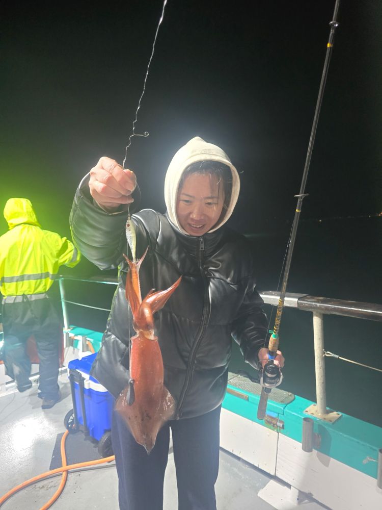 Person holding a squid on a fishing line at night on a boat, wearing a hooded jacket.