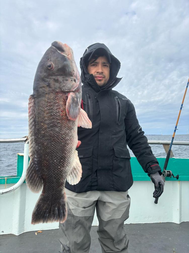 Person in a black jacket holding a large fish on a boat with a cloudy sky.