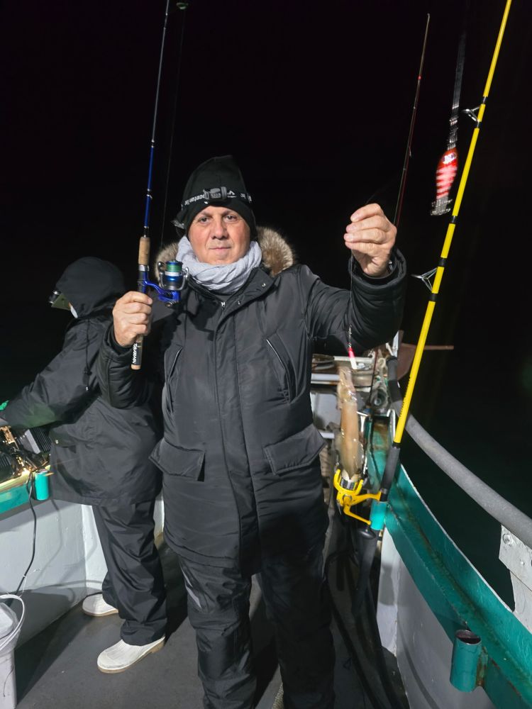 Person in winter clothing fishing at night on a boat, holding a fishing rod.