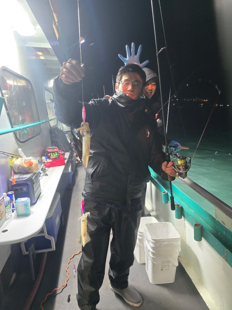 Two people on a boat at night, one holding a fishing rod with squid, another holding a blue glove above his head.