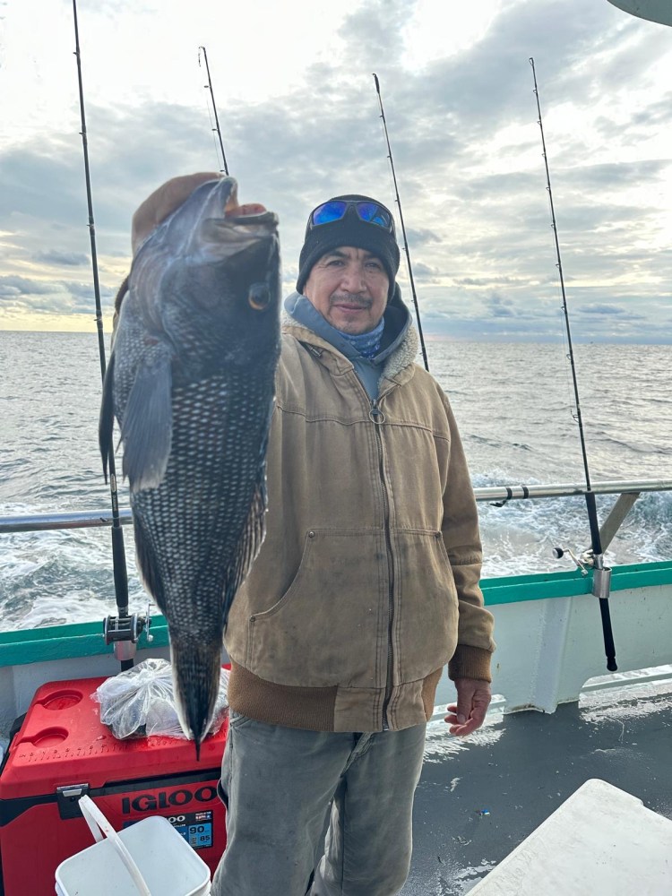 Person in a brown jacket holding up a large fish on a boat with ocean in the background.