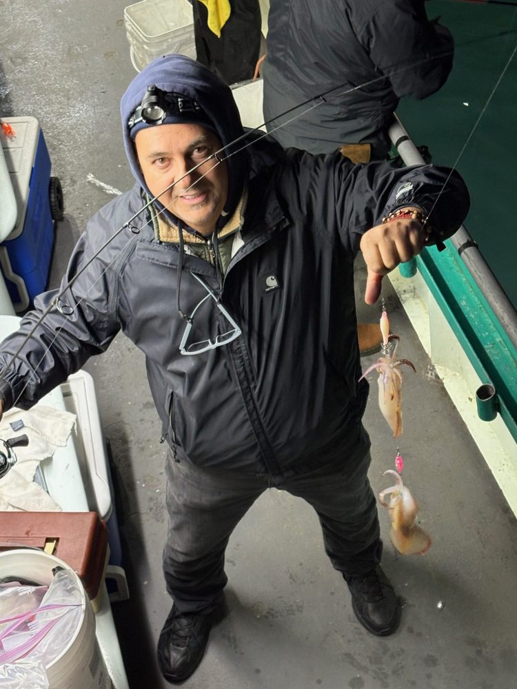 Person on a boat holding up two caught squids on a fishing line, wearing a headlamp and a black jacket.