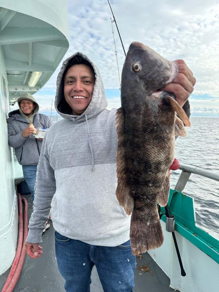 Smiling person on a boat holding a large fish, another person in the background with a sandwich.