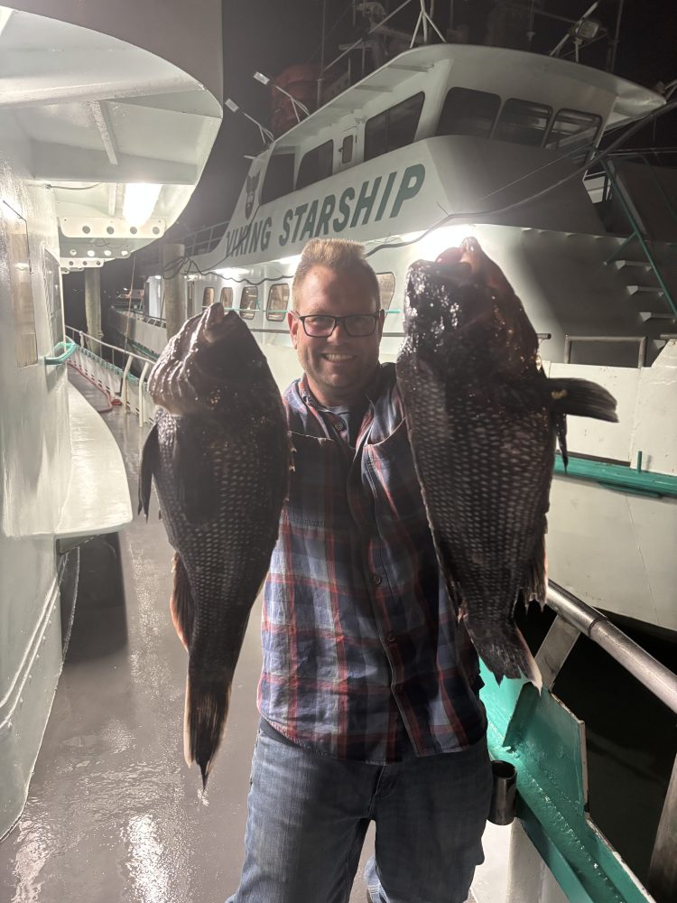 Person holding two large fish on a boat deck at night.