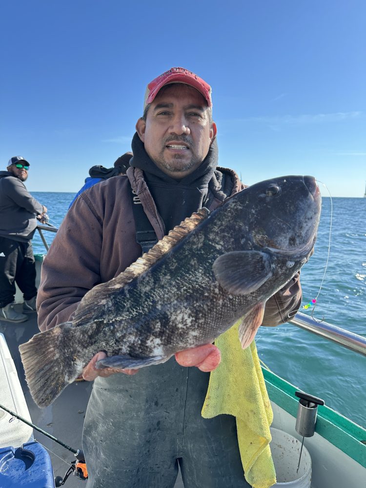 Man holding large fish on a boat, with another person fishing in the background.