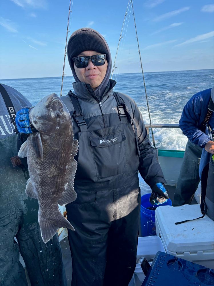 Person holding a large fish on a boat, wearing sunglasses and warm clothing.