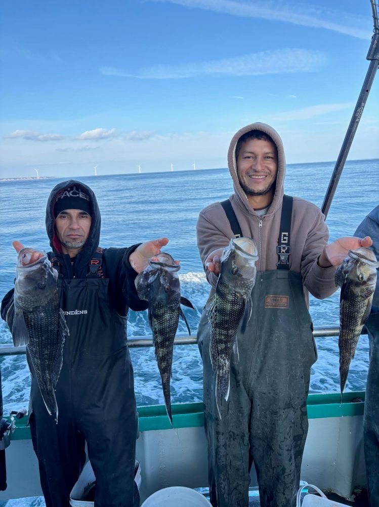 Two people on a boat holding freshly caught fish with sea in the background.