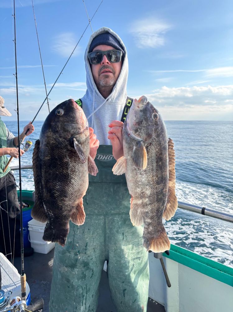 Person holding two large fish on a boat at sea, wearing a hoodie and sunglasses.
