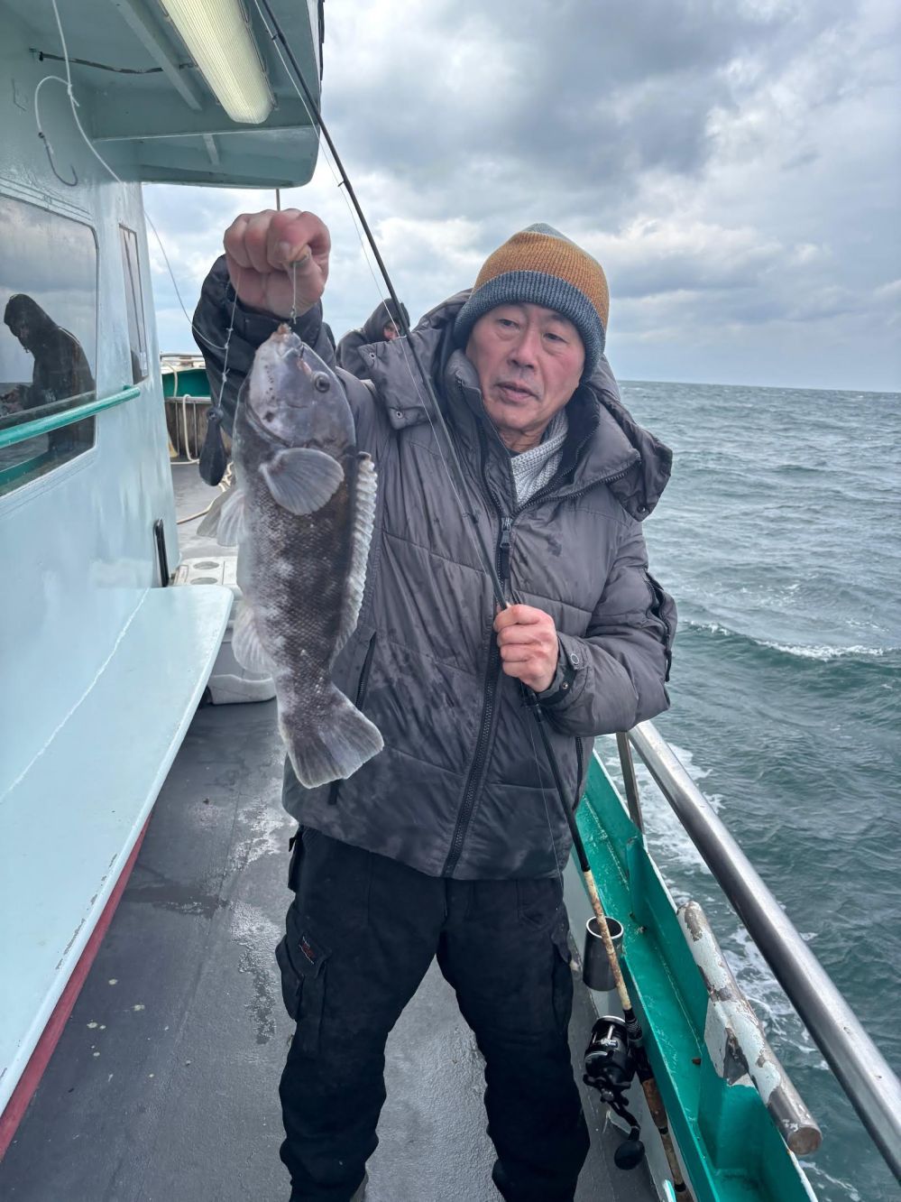 Person in a winter jacket holding a fish on a boat with a cloudy sky in the background.