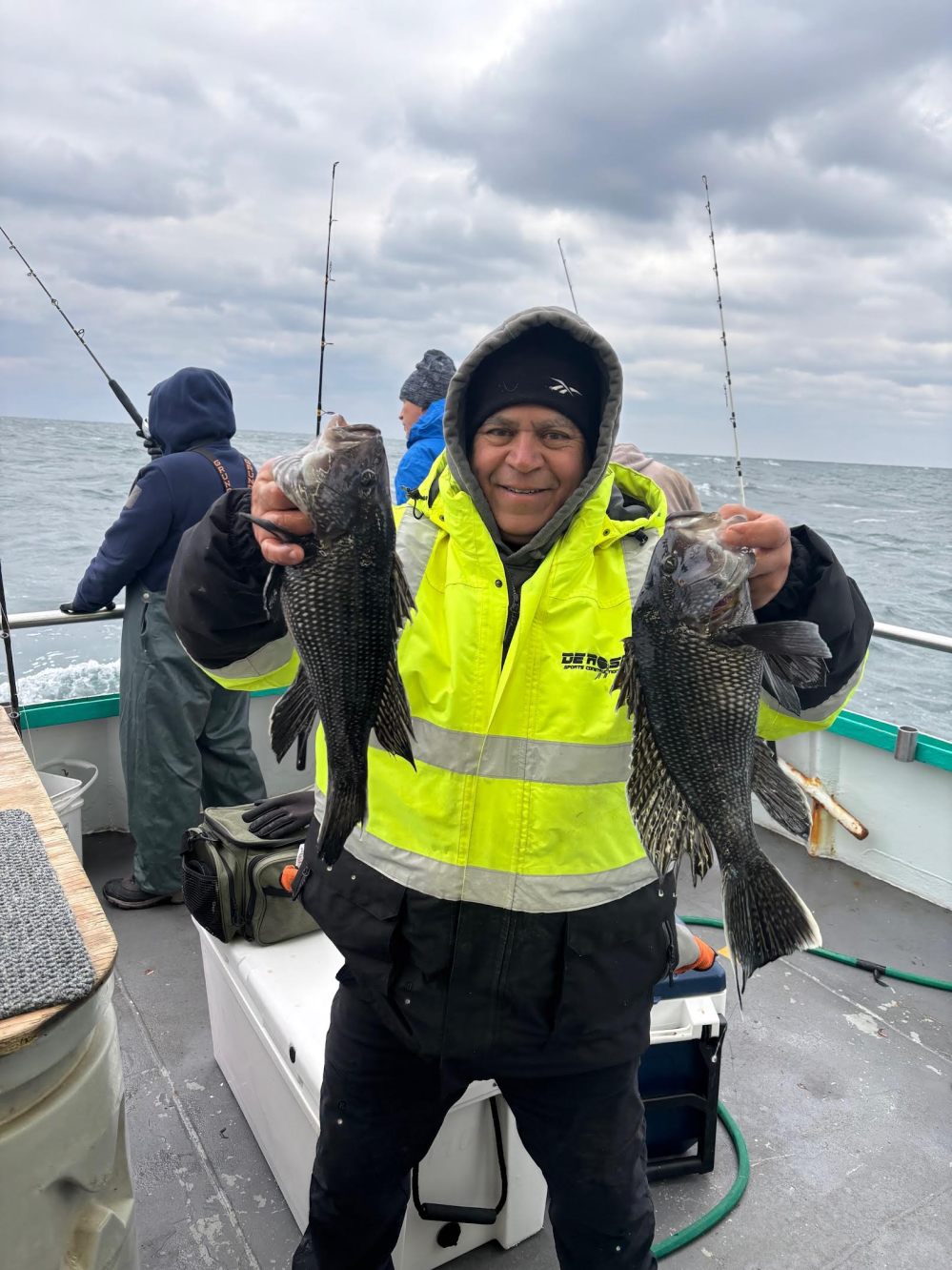 Person in yellow jacket holding two fish on a boat with cloudy sky.