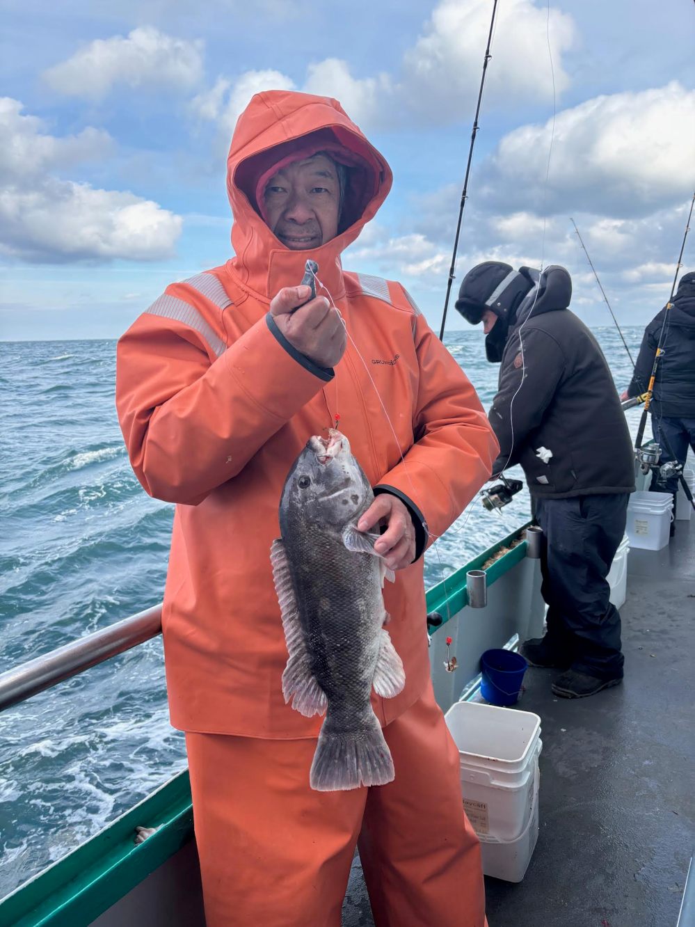 Person in orange raincoat holding a fish on a boat with another person fishing nearby.
