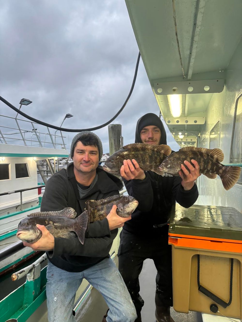 Two people on a boat holding three large fish under a cloudy sky.