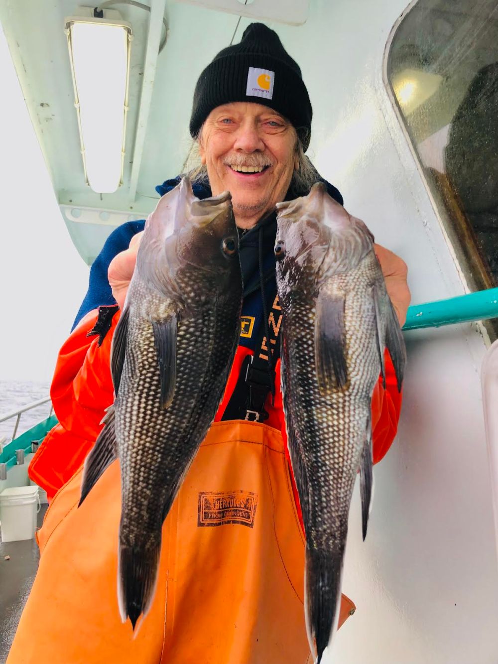 Person in orange bib holding two large fish on a boat, smiling and wearing a black beanie.