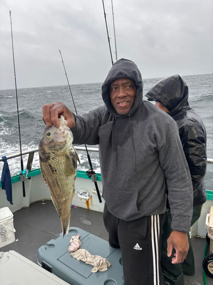 Man in hoodie on boat holding a fish, with another person in a hooded raincoat, cloudy sky and ocean in background.