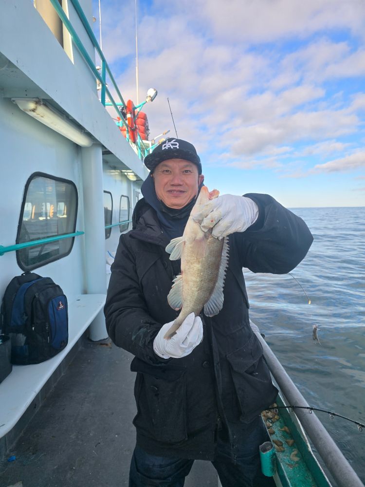 Person on a boat holding a fish, wearing a cap and gloves, with ocean and cloudy sky in the background.