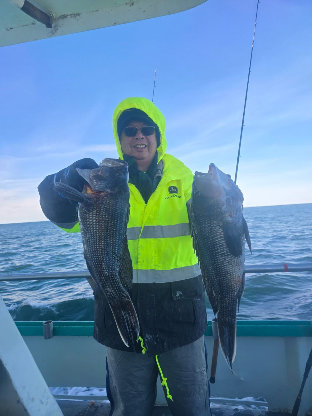 Person in yellow jacket holding two fish on a boat with ocean in background.