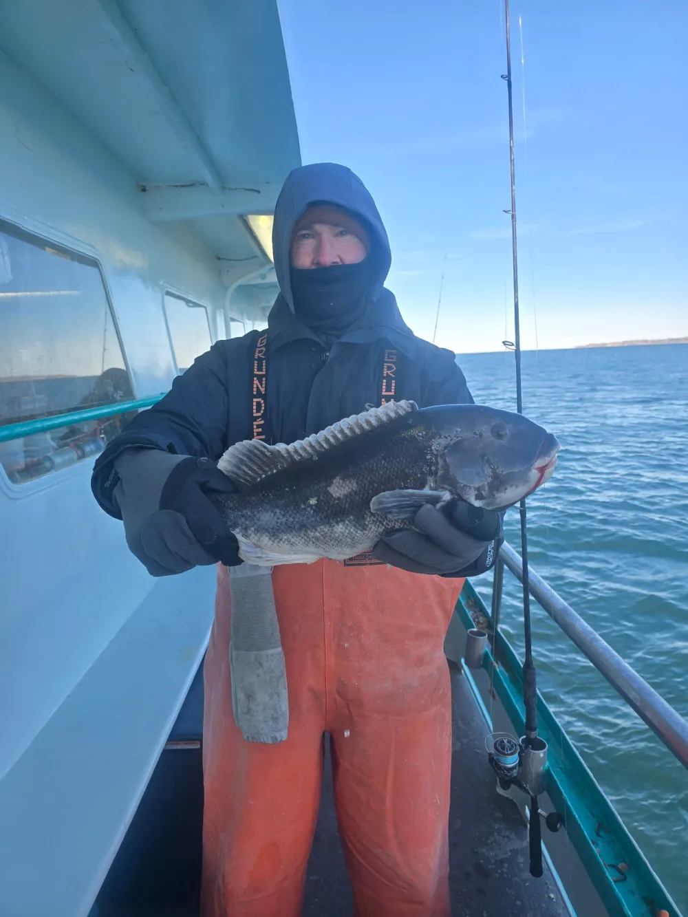 Person in winter gear holding a large fish on a boat with ocean in the background.