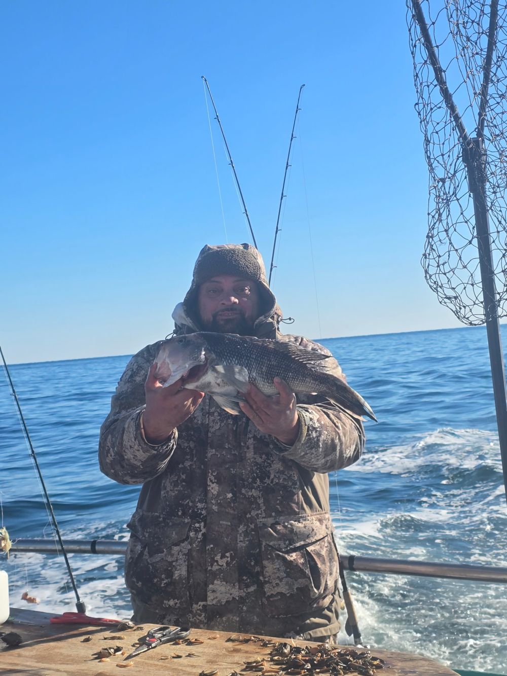 Person in camo holds a fish on a boat with fishing gear and ocean background.